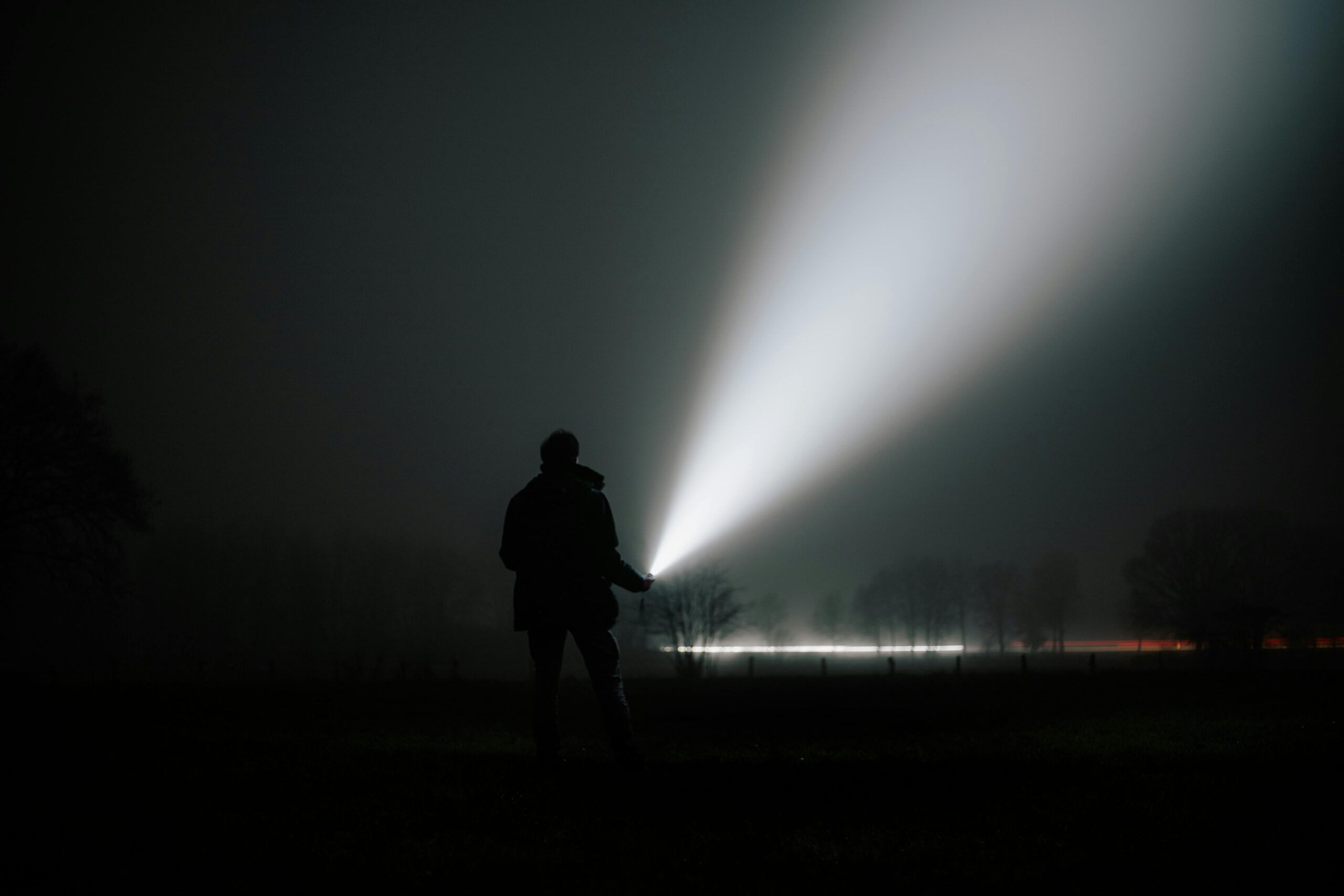 A person holds a flashlight in a foggy night landscape, creating dramatic light trails.